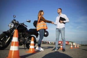 A woman in a safety vest shaking hands with an instructor at a motorcycle training course, with traffic cones and a motorcycle in the background.